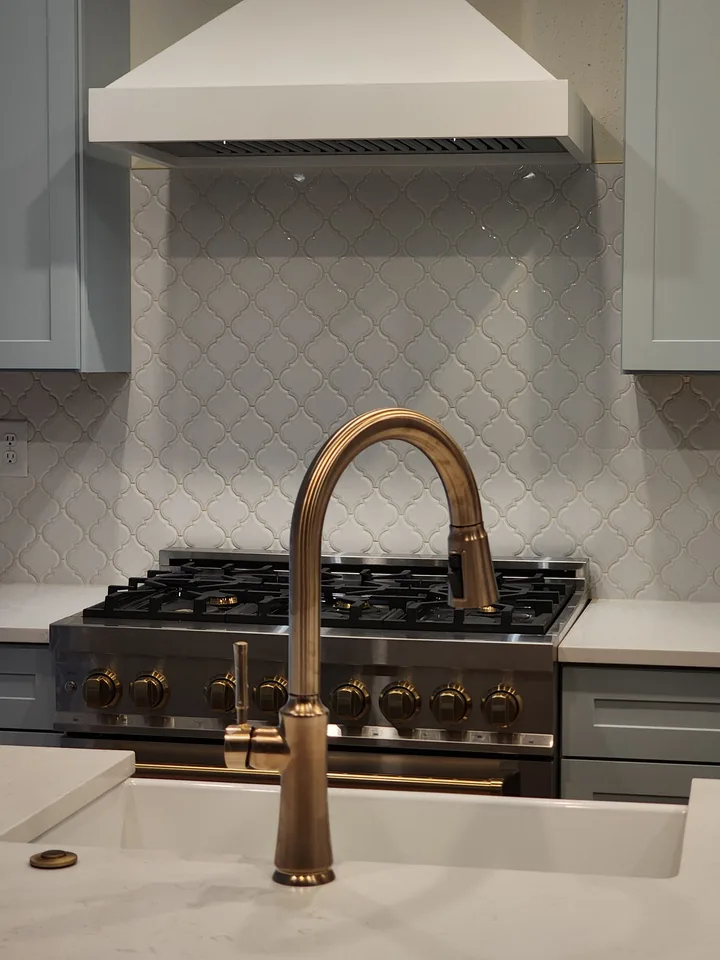 Close view of a white arabesque-pattern range wall with pale blue cabinets and warm-metal fixtures