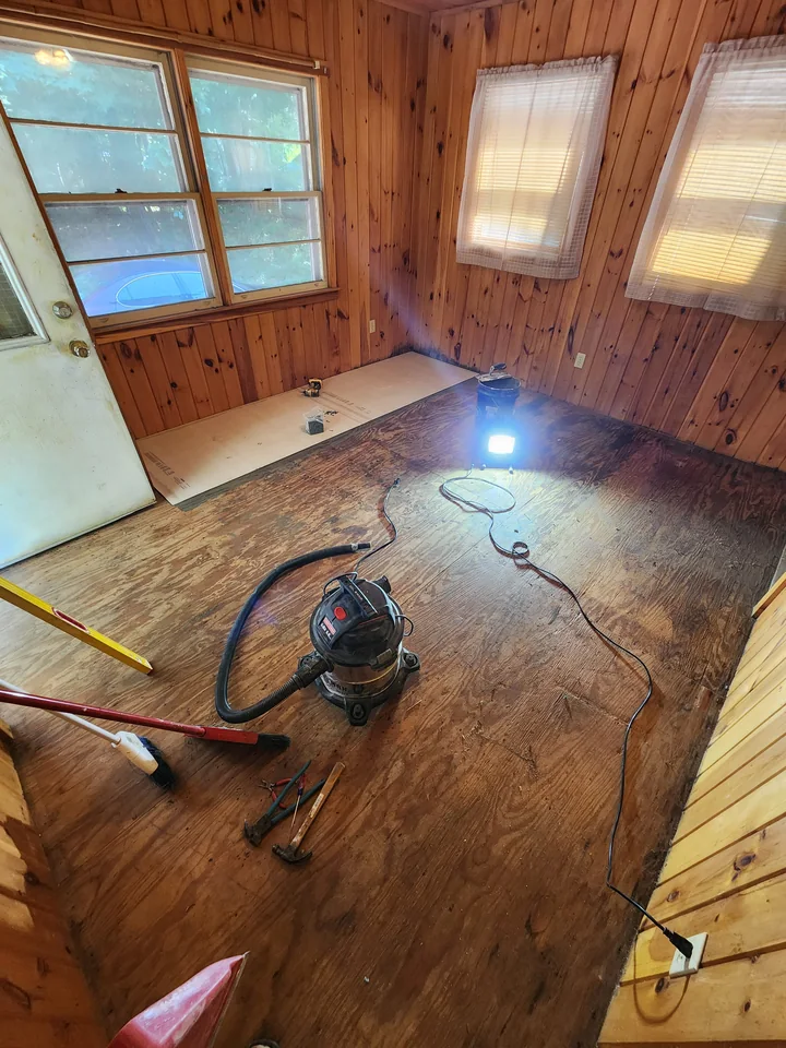 Wood-paneled room with the original floor exposed and the first section of underlayment in place