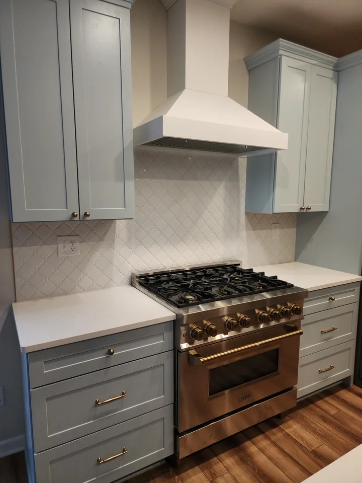 White arabesque-pattern backsplash behind a range, framed by pale blue cabinets and a white hood